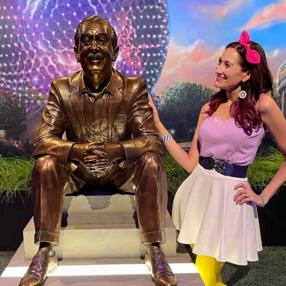 Theresa from the No-Guilt Disney Podcast posing next to the Walt the Dreamer statue, which features him sitting with his hands clasped together and arms resting on his knees.