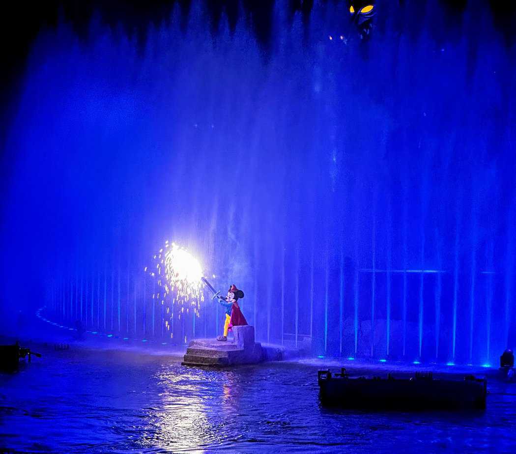 Fantasmic Disney World: Is it too scary for kids (2023) Parents guide. Mickey Mouse holding a sword with sparks coming off the end in front of water fountains spraying into the sky.
