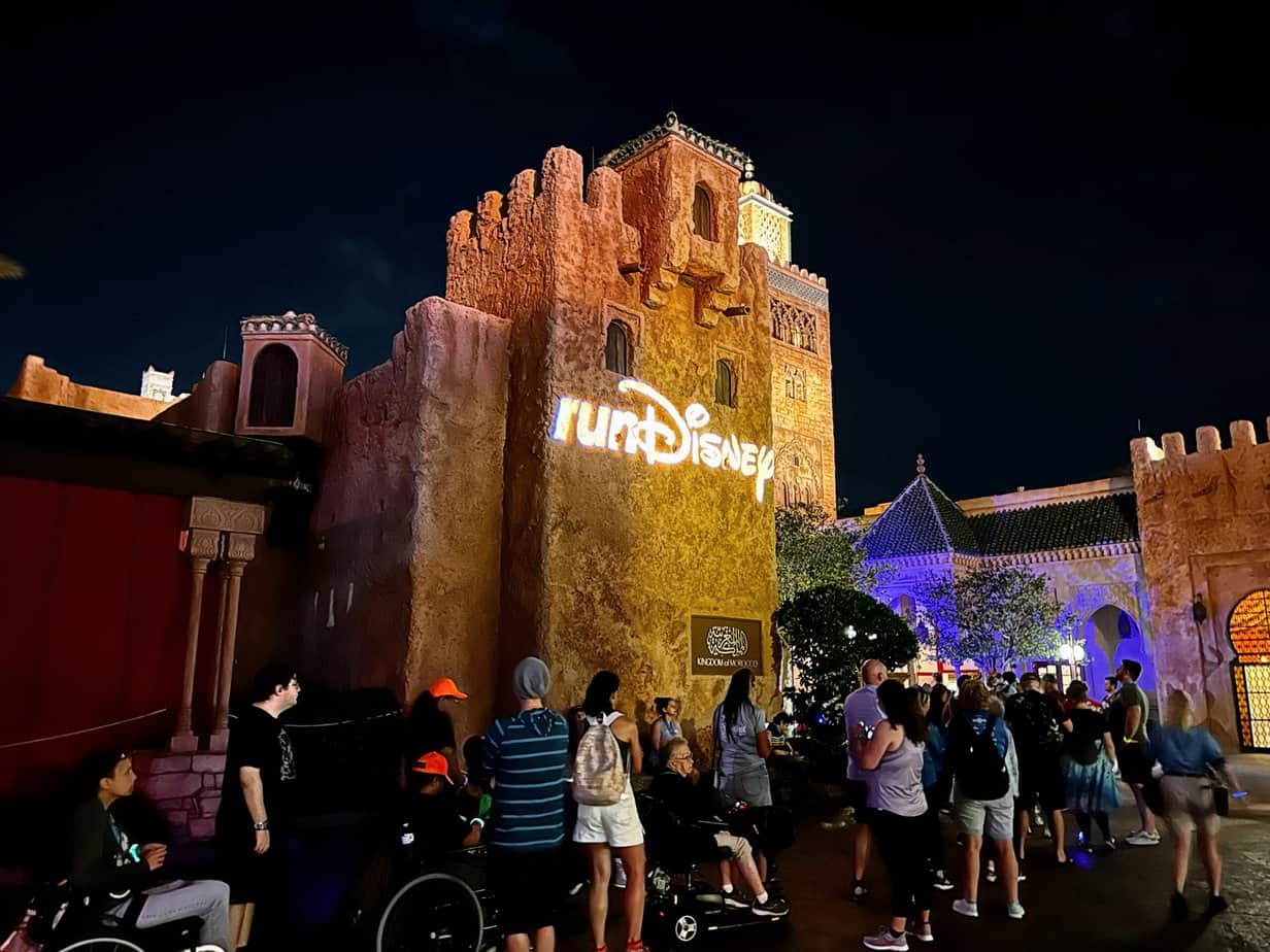 rundisney wine and dine race weekend has an after party held in Epcot for runners and guests. This exclusive party only happens at the wine and dine race. people standing in line to meet a character in front of the Morocco Pavillion which has a runDisney sign on it.